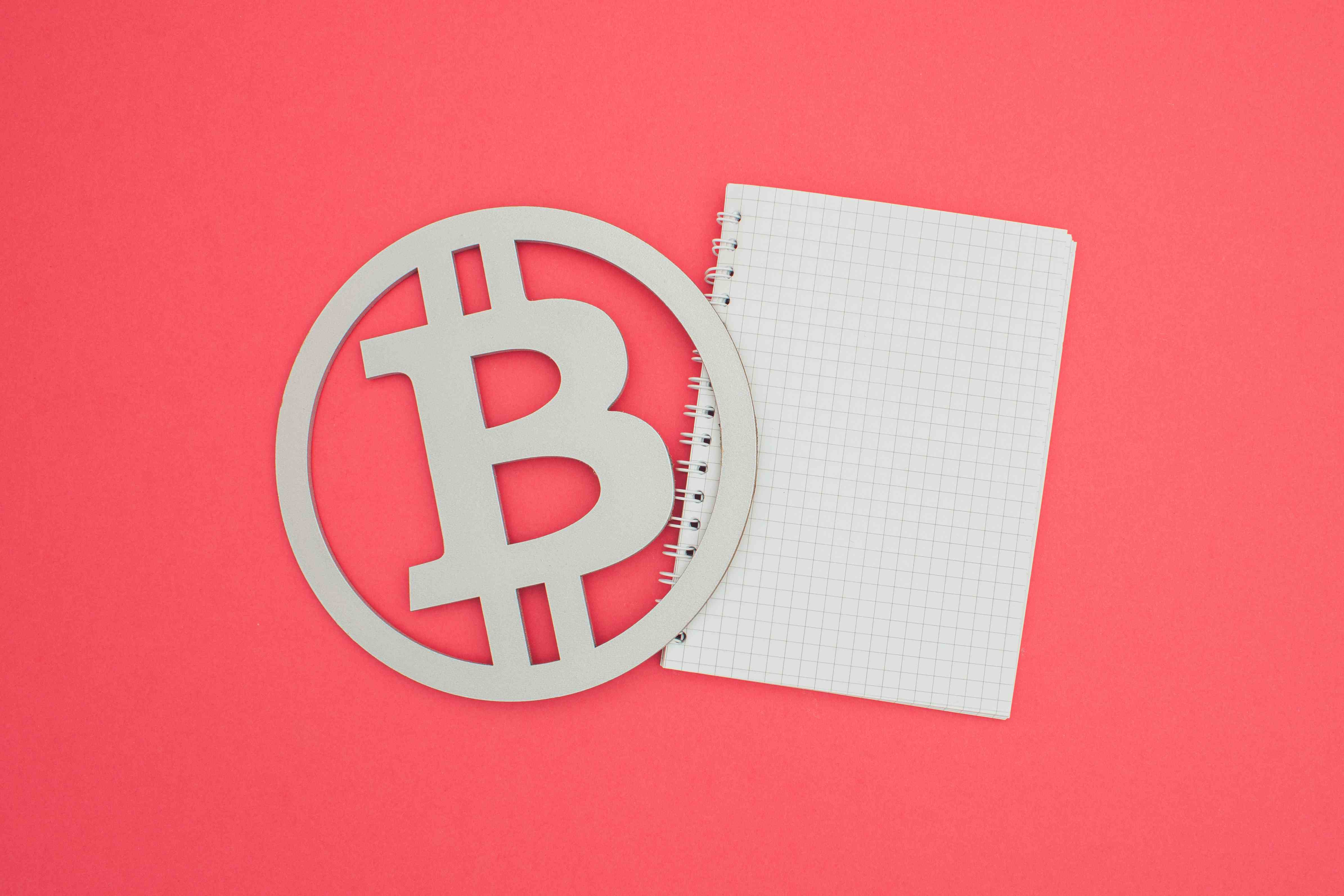 Bild eines Bitcoin-Symbols und eines weißen Notizbuchs vor einem lachsfarbenen Hintergrund.