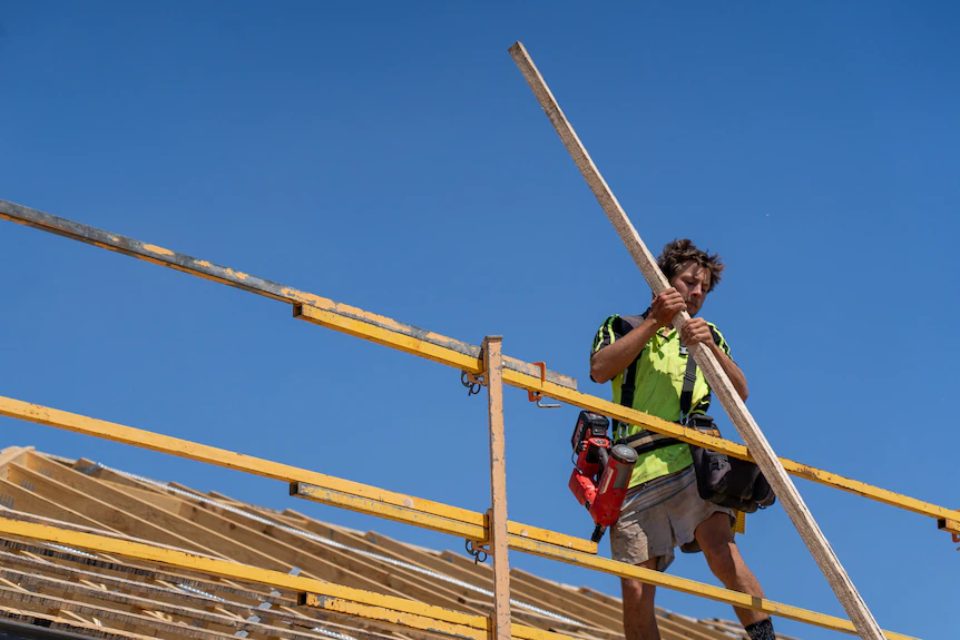 Un homme sur un toit soulève une planche de bois au-dessus d'un échafaudage. 