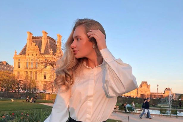 Une jeune femme blonde se brosse les cheveux en arrière debout dans une rue de Paris dans une blouse blanche et un pantalon en cuir
