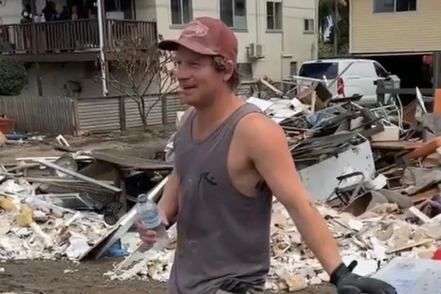 Homme en casquette en maillot de corps appuyé sur une remorque avec une bouteille d'eau à la main devant les décombres 