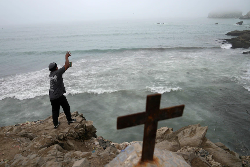 Une croix dépasse de la terre tandis qu'en arrière-plan un homme jette sa ligne de pêche des rochers dans l'océan gris.