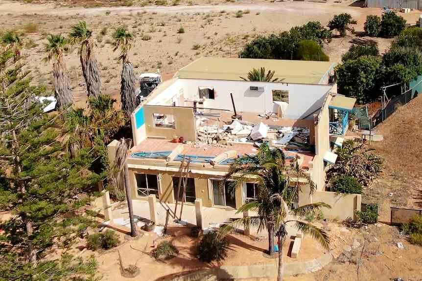 Une maison de deux étages sans toit et des dommages au dernier étage couvert de débris.  Un terrain de sable ouvert entoure la maison.