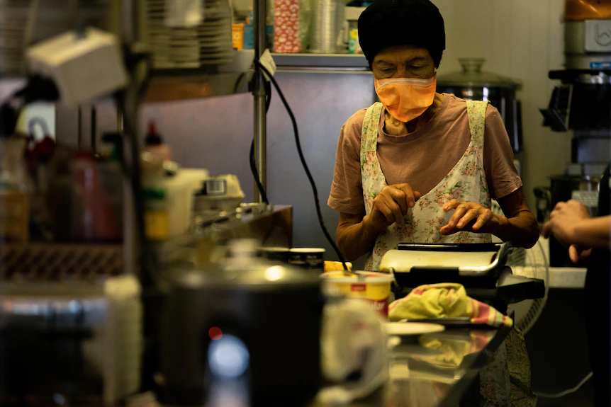 Une femme avec un masque et un tablier dans une petite cuisine de cantine.