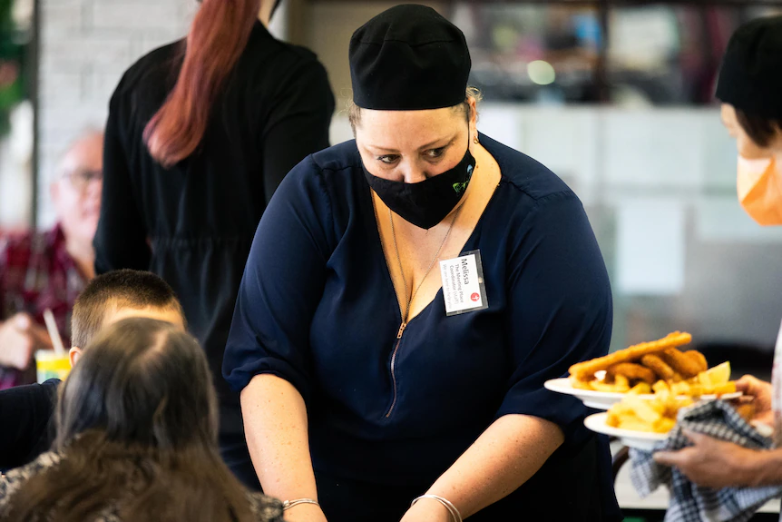 Une femme portant un masque sert des clients à une table dans un café.