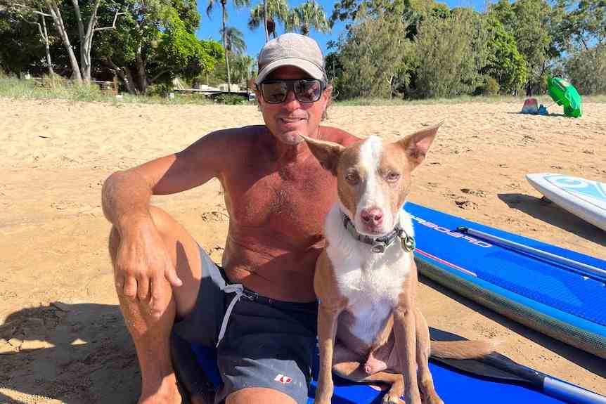 Un homme souriant avec une casquette et des lunettes est assis torse nu sur un paddleboard avec son chien à côté de lui.