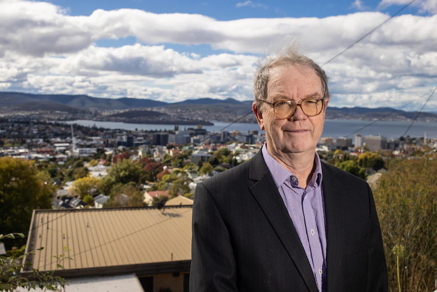 Un homme portant des lunettes et un costume avec la ville de Hobart en arrière-plan.