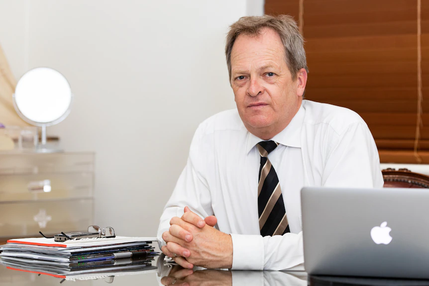 Un homme en chemise blanche avec une cravate noire et dorée regarde la caméra assis à un bureau avec un plateau en verre.