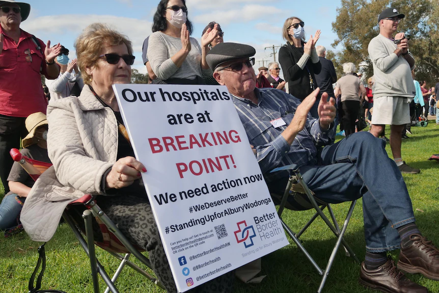 Un homme et une femme sont assis sur des sièges de pique-nique lors d'un rassemblement et une femme brandit une pancarte avertissant que les hôpitaux sont sous pression.