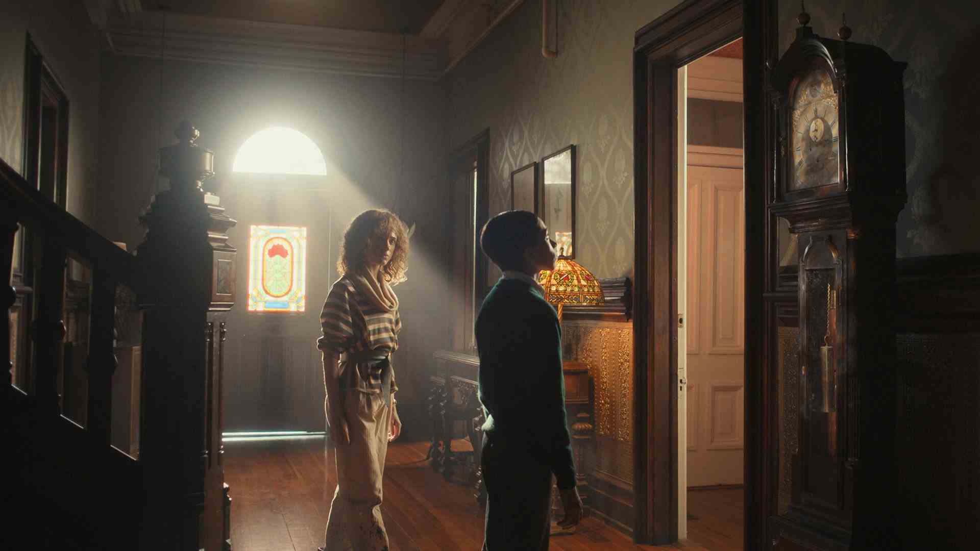 Dans le hall d'une vieille maison, un garçon regarde une horloge grand-père tandis qu'une femme se tient debout et le regarde.