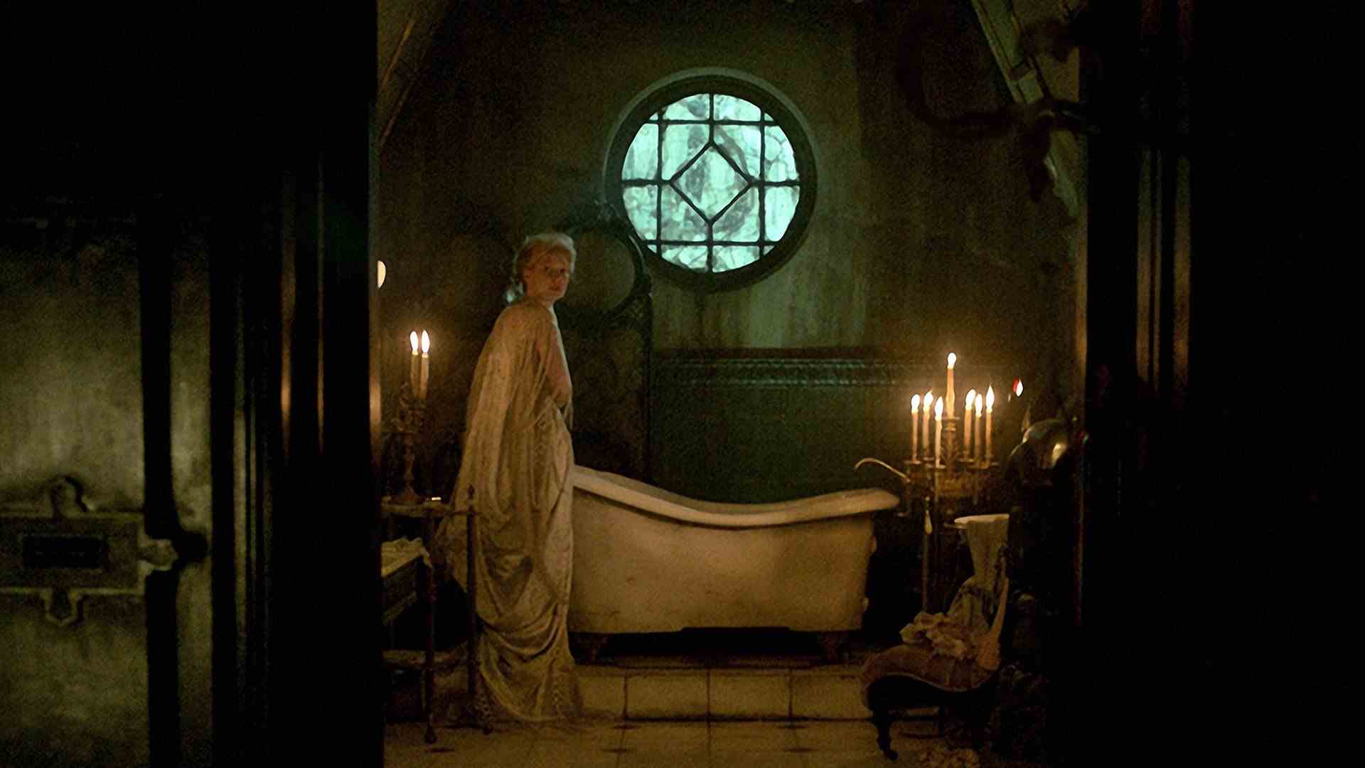 Une femme se tient près d'une baignoire sur pattes sous une fenêtre ronde.