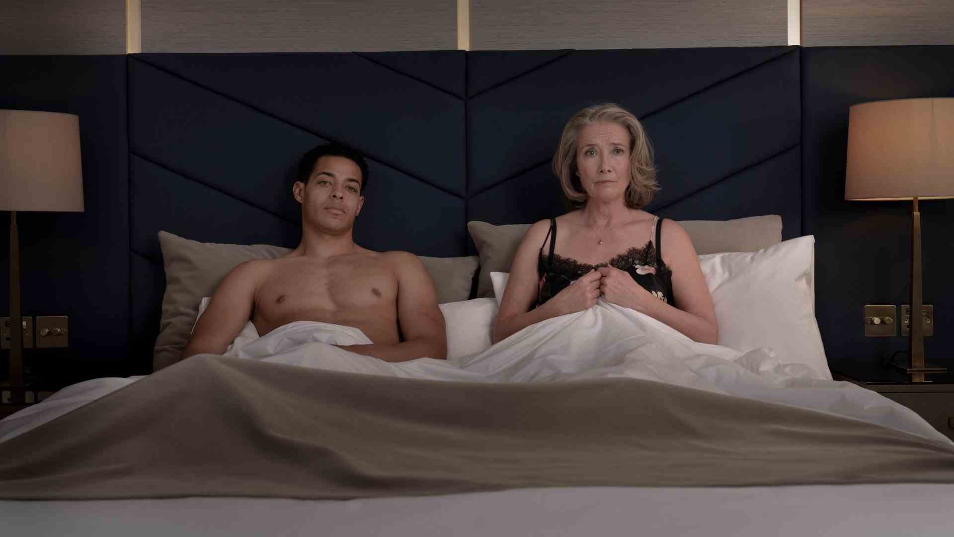 Un homme et une femme en sous-vêtements sont assis dans leur lit, les couvertures relevées et regardent la caméra.