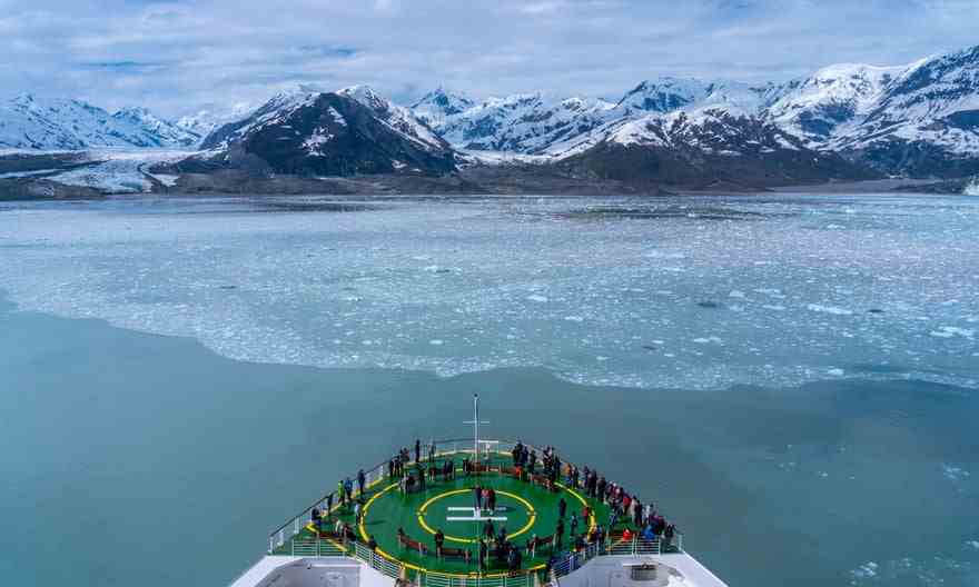 Bateau de croisière naviguant vers un glacier, avec stand de passage sur l'héliport du navire, surplombant un iceberg géant.
