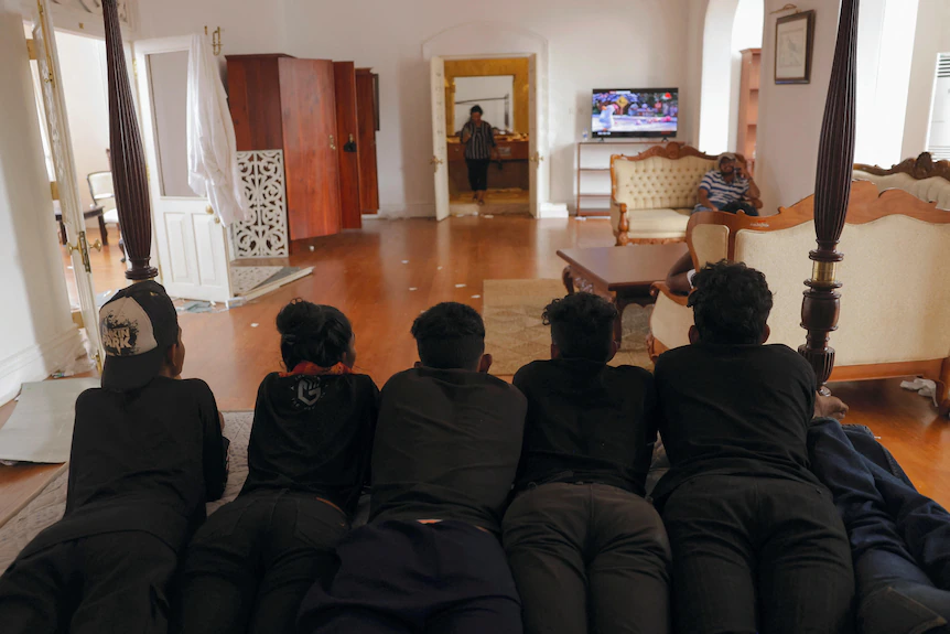 Des hommes vêtus de noir sont allongés face contre terre sur le lit du président et regardent le cricket sur une télévision à proximité. 