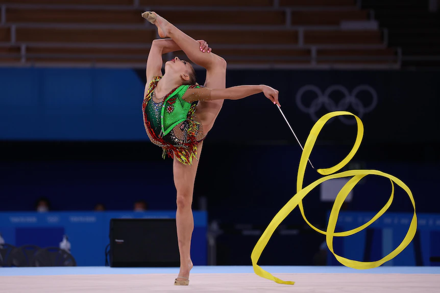 Une gymnaste rythmique se tient sur la pointe des pieds tout en tenant son autre jambe derrière son dos et en agitant un ruban. 