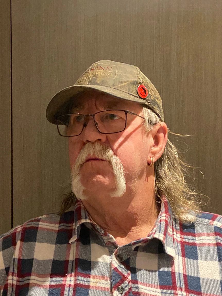 Un vieil homme avec une grande moustache portant une chemise à carreaux et une casquette.