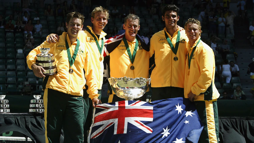 Une équipe nationale de tennis australienne se tient à côté de la Coupe Davis drapée du drapeau australien.